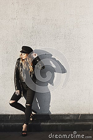 Modish blonde model girl in a leather jacket, ripped jeans, and black cap standing outdoors against the urban wall in Stock Photo