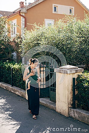 Mom with a baby on a sling walks down the street past the fence of a house with greenery Stock Photo