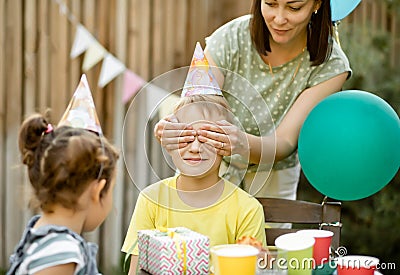 Mom from behind closes eyes her son with hands. Cute funny nine year old boy celebrating his birthday with family and friends with Stock Photo