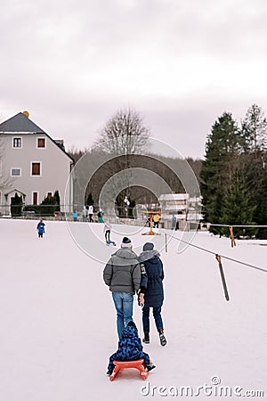 Mom and dad are carrying a small child on a sled climbing a hill. Back view Stock Photo