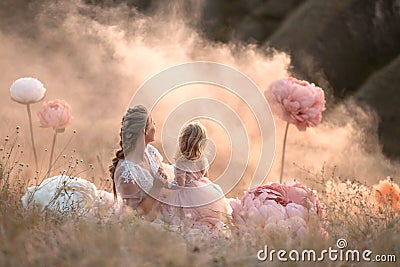 Mom and daughter in pink fabulous dresses look into the distance, sitting in a field surrounded by large pink decorative flowers Stock Photo