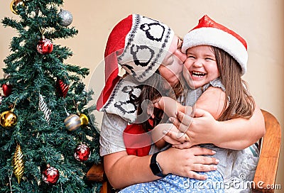 Mom and daughter in the red santa hat near Christmas tree. Mom kisses the child Stock Photo