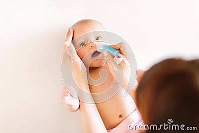 Mom holding her head to a small girl with her hands sucks the snot from the baby`s nose lying on the bed through the Stock Photo