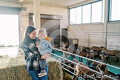 Mom with a little girl in her arms stands by a haystack near a goat paddock Stock Photo