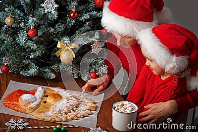 Mom tells her daughter a story about Santa next to a Christmas tree and a cake in the form of Santa Claus Stock Photo