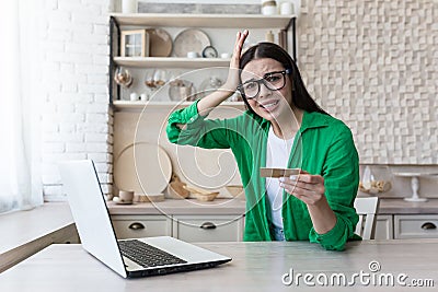 Monetary crisis. Credit debt. Upset young woman holding credit card, sitting at laptop at home Stock Photo