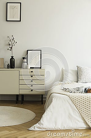A monochromatic beige and white bedroom interior with a view at a bed and a drawer cabinet standing on a wooden floor. Real photo. Stock Photo