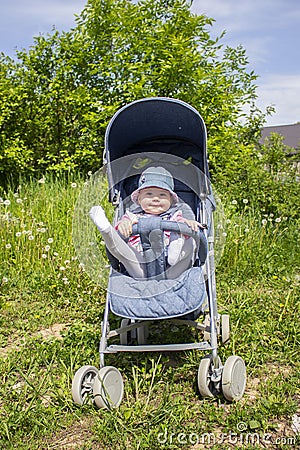 A 9 month old girl sits in a summer carriage and plays with her legs. Little smiling child in a hat on a walk, summer day Stock Photo