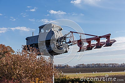 Monument to old caterpillar tractor with mounted plough against field Editorial Stock Photo