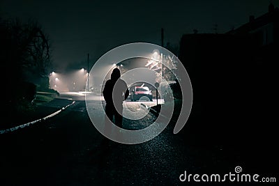 A moody concept of a scary hooded figure. Standing in the middle of the road in a quiet suburban street. On a foggy winters night Stock Photo