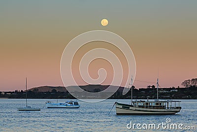 A full moon rising above boats in a harbor Stock Photo