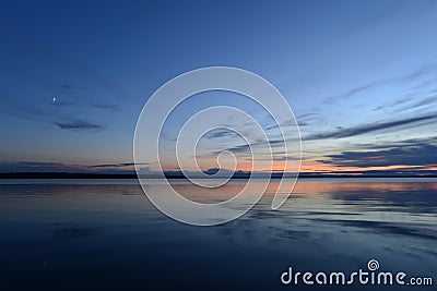 Moonrise in twilight light in the blue sky at sunset over the quiet mirror water of the lake Stock Photo