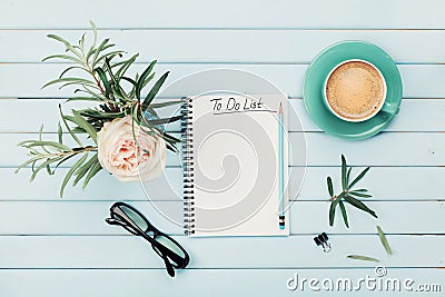 Morning coffee cup, notebook with to do list, pencil, eyeglasses and vintage rose flower in vase on blue rustic table from above. Stock Photo