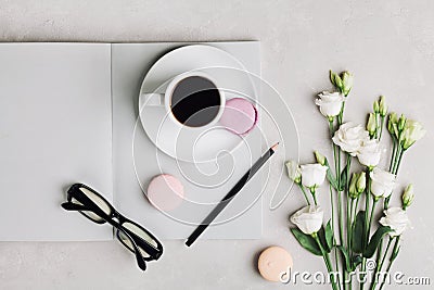 Morning cup of coffee, empty notebook, pencil, glasses, white flowers and cake macaron on light table top view. Stock Photo