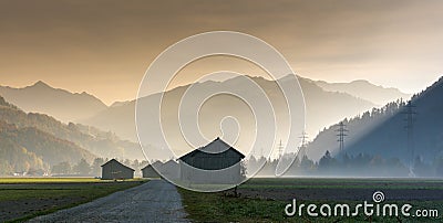 Morning mist in a mountain valley with fields and old wooden barns and lattice cross power lines and mountains in silhouette behin Stock Photo