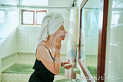 Woman in towel applies sunscreen to face in vintage bathroom, protecting skin against UV rays. Morning skincare routine Stock Photo