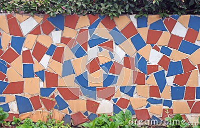 Mosaic of large broken colorful tiles covering a garden wall with plants along the top and bottom Editorial Stock Photo