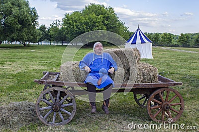 A contented man in medieval blue clothes is sitting on a horse-drawn cart on the background of a Editorial Stock Photo