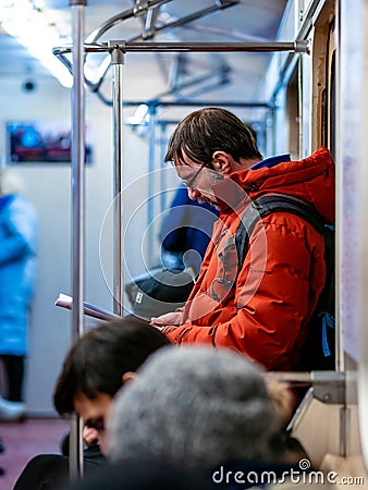 Moscow, Russia - February 8, 2020: Man rides in subway car and reads a magazine. Guy with glasses and orange jacket is standing at Editorial Stock Photo