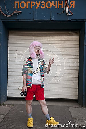 A young man in a motley Hawaiian-style shirt, white T-shirt, red shorts and a pink fur hat with a camera stands in front of the Editorial Stock Photo