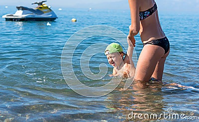 Mother in black sunglasses and smiling baby boy son in green baseball cap playing in the sea in the day time. Positive human emot Stock Photo