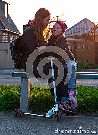 Mother and daughter on a bench in the park. Child riding on kickscooter. Blonde cute girl on scooter spring city street Stock Photo