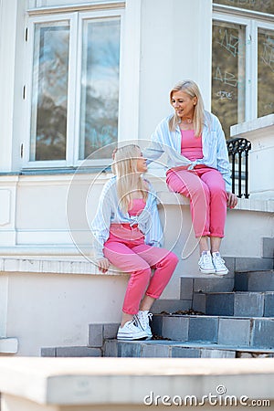 Mother and daughter sitting near stairs and laughing. Mom straighten white hair of girl. Funny talk between relatives. Stock Photo