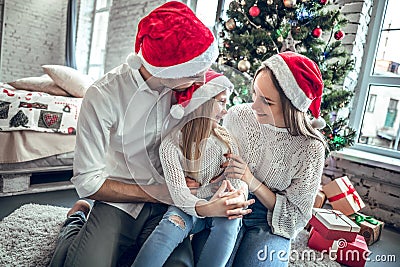 Mother, father and daughter with attractive appearance look at each other`s eyes, sit on white carpet near decorated New Year tree Stock Photo