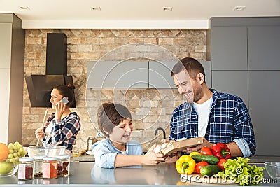 Family at home in kitchen together father and son standing looking at eggs laughing playful while mother holding phone Stock Photo