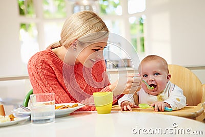 Mother Feeding Baby Sitting In High Chair At Mealtime Stock Photo