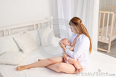 Mother feeds baby 6 months of breast sitting on a white bed at home, place for text Stock Photo