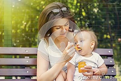 Mother giving her infant child complementary feeding pumpkin puree in sunny day outdoor. Both mom and kid sit on bench in park Stock Photo