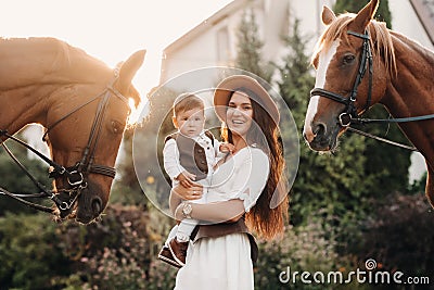 A mother in a hat with her son in her arms stands next to two beautiful horses in nature. a family with a child is photographed Stock Photo