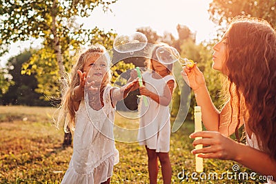 Mother helps daughters to blow bubbles in summer park. Kids having fun playing and catching bubbles outdoors Stock Photo