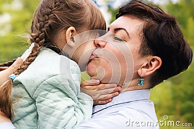 A mother with her eyes closed, kisses her little daughter on the cheek in the open air. A little daughter hugs her mother by the Stock Photo