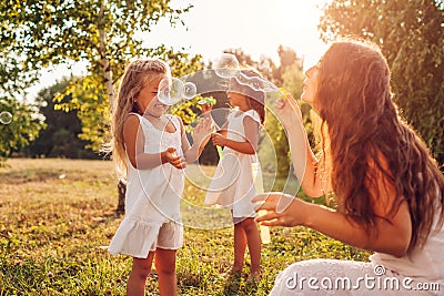 Mother`s day. Woman helps daughters to blow soap bubbles in summer park. Kids having fun playing outdoors Stock Photo