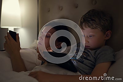 Mother with son looks in their electronic devices lying in bed Stock Photo