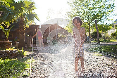 Mother sprays a child with a hose in the courtyard of the house, Boy drenched in water on a hot sunny day Stock Photo