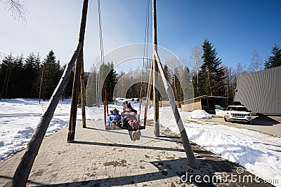 Mother swinging with daughters in big wooden swing in early spring snowy mountains against tiny house and car Stock Photo