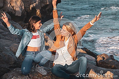 Mother and daughter sitting on the rocks by the mediterranean sea with arms raised playing with the air Stock Photo