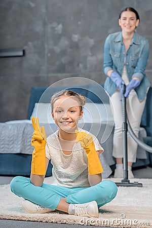 Mother with vacuum cleaner looking at happy daughter in rubber gloves gesturing Stock Photo