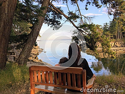 A mother and young daughter sitting side by side on a bench overlooking the ocean on a beautiful evening in the gulf islands Stock Photo