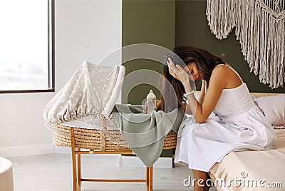 Young tired depressed mother holding milk bottle sitting near bassinet with newborn baby at home Stock Photo