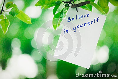 Motivating phrase Believe in yourself. On a green background on a branch is a white paper with a motivating phrase. Stock Photo