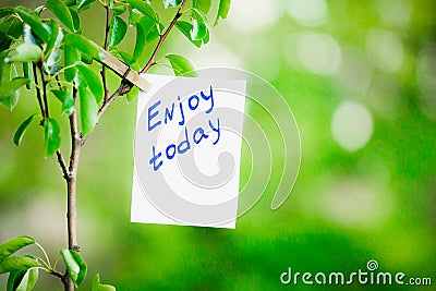 Motivating phrase enjoy today. On a green background on a branch is a white paper with a motivating phrase. Stock Photo