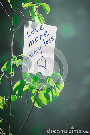 Motivating phrase love more worry less. On a green background on a branch is a white paper with a motivating phrase Stock Photo