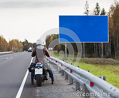 Motorcyclist standing on the highway in front of the blank information board with blue background, autumn season Stock Photo