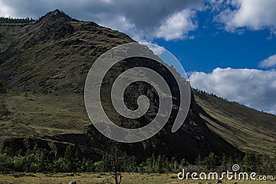 Mountain on Baikal, Sarma. Large tall green grassy round rock. Trees and bushes in the foreground in dry steppe. Cloudy blue sky Stock Photo