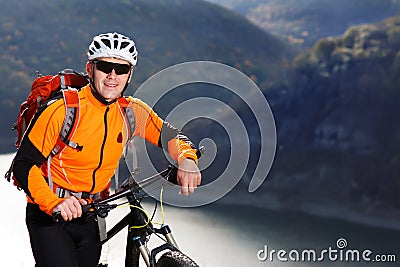 Mountain cyclist in the orange jacket stands under the beautiful river against mountains. Stock Photo