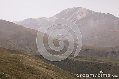 Mountain landscape - mountain peak with glacier in soft light white dim mist, yellow hilly folded slopes with dry alpine meadow Stock Photo